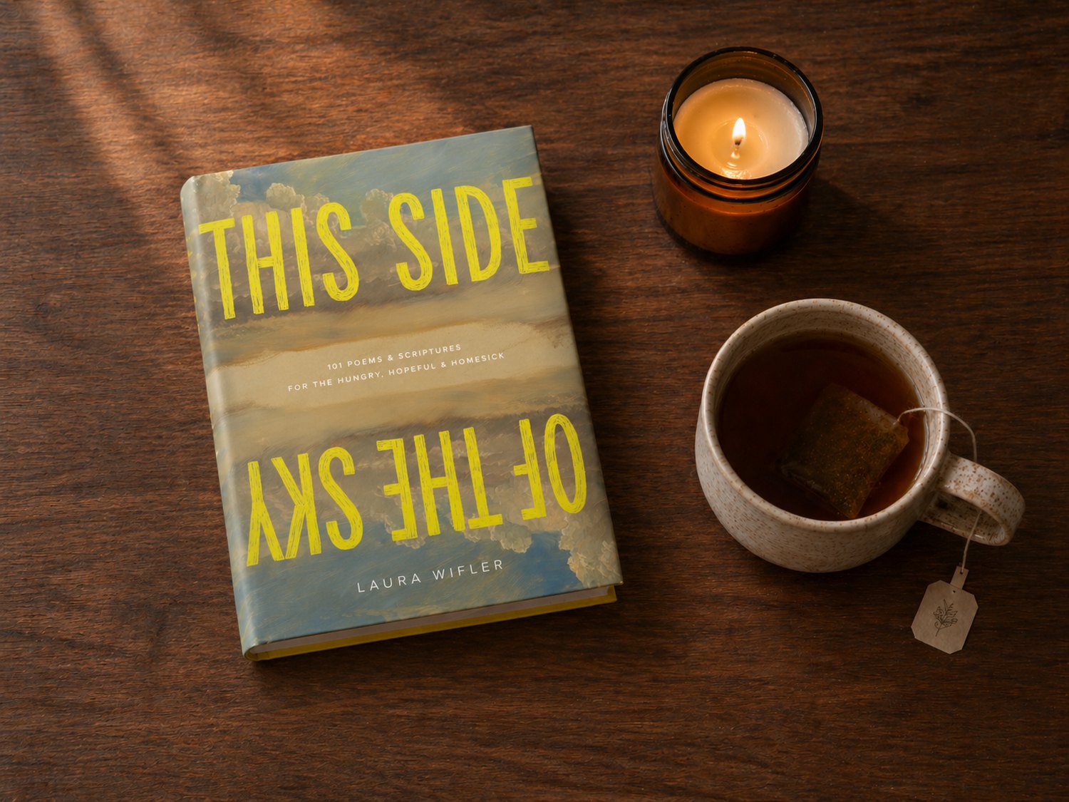 This Side of the Sky on a wood table beside a lit candle and a cup of tea